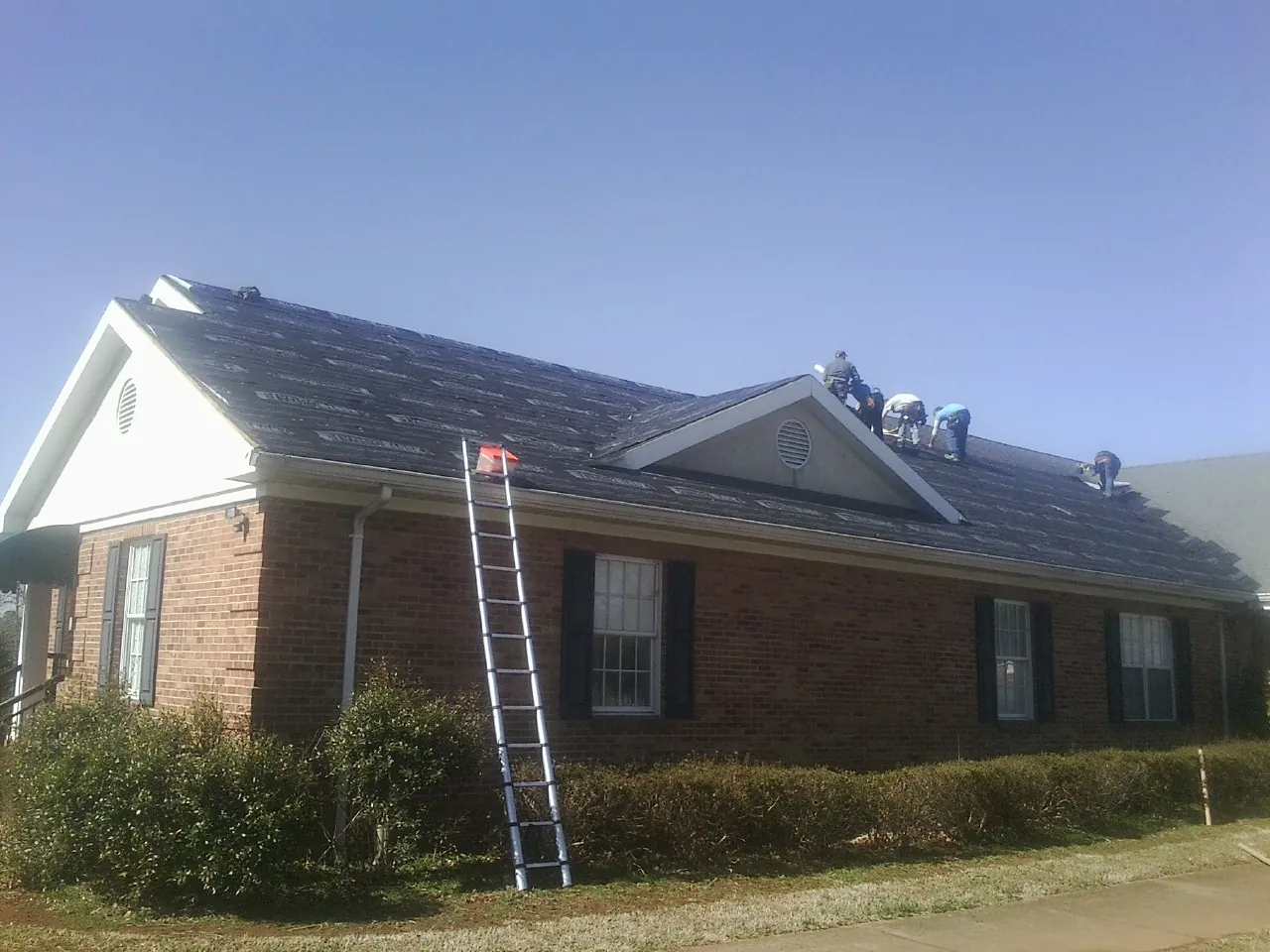 Crew tearing shingles off a brick ranch roof with a ladder set — Emergency Roof Repair work by our crew in Goshen